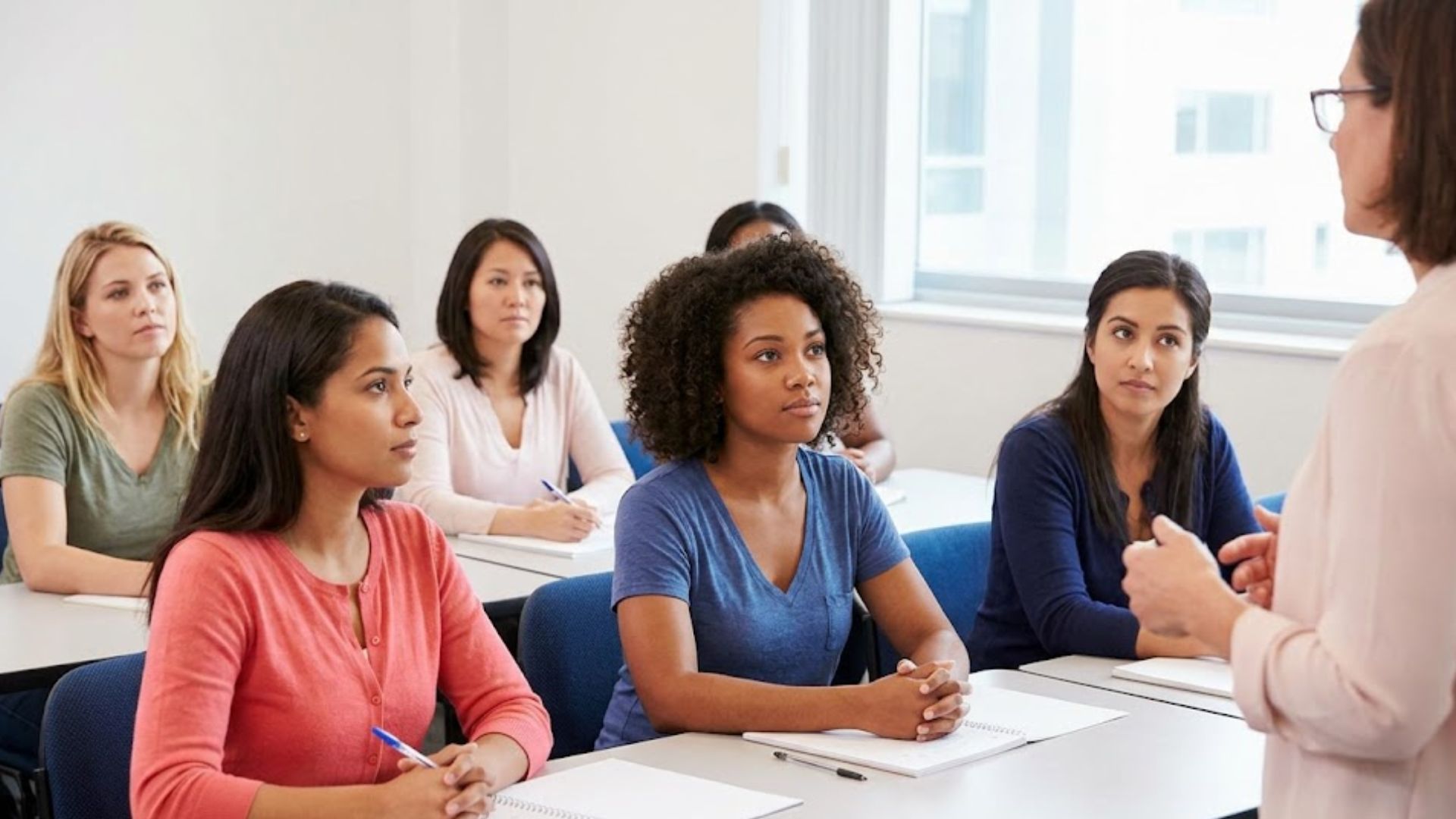 Diverse women in a classroom, intently listening to an instructor.